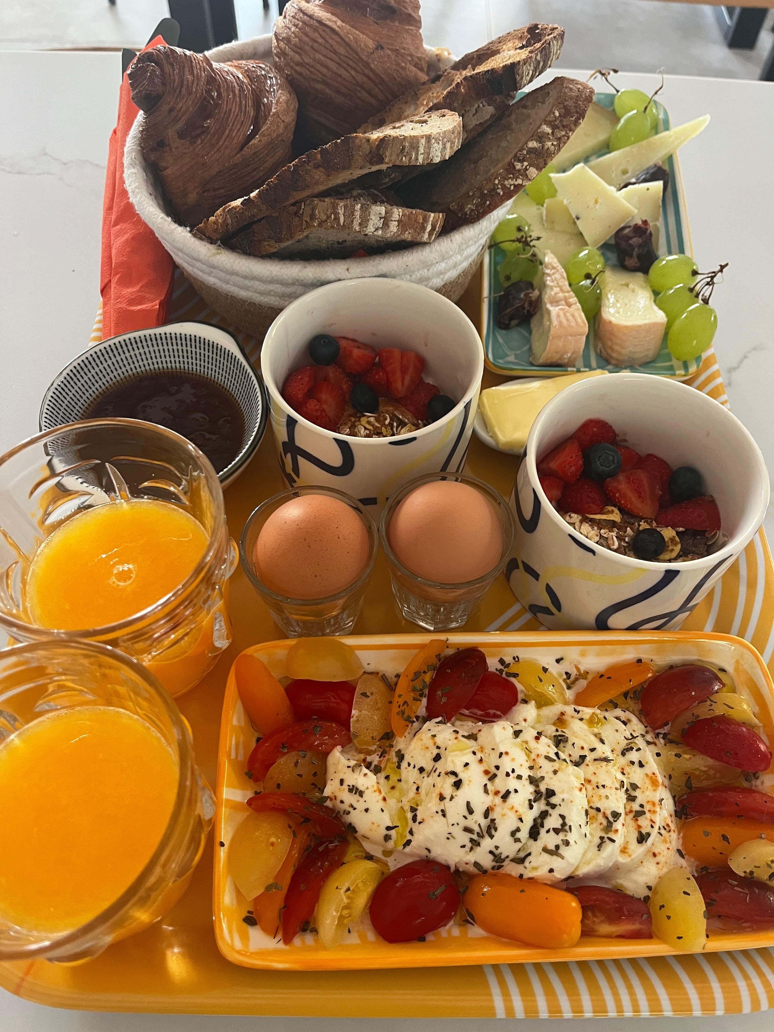 Breakfast tray: pastries, country bread, cheeses and grapes, yoghurts with red fruit and granola, eggs, burrata with heirloom tomatoes and orange juice